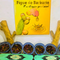Painting of a Moroccan display showcasing prickly pear (figue de barbarie) products. A bright yellow sign with a cactus and dripping oil hangs above bowls filled with dark seeds, set on a green patterned cloth. Natural wood branches frame the scene, highlighting the organic and traditional feel. The following images show the same artwork displayed in different interior settings.
