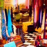 Painting of a colourful weaving studio filled with handmade rugs, yarn, and a loom in progress. Bright reds, blues, yellows, and earth tones fill the scene, with patterned textiles hanging on the walls and a woven basket of tools on the floor.The following images show the same artwork displayed in different interior settings.