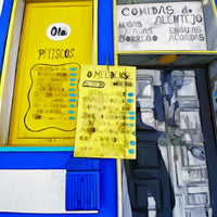 Painting of a Portuguese café window with bright yellow and blue trim, handwritten yellow menus displaying traditional Alentejo dishes like migas, favas, and borrego. A white sign above the door reads “Comidas do Alentejo.” The following images show the same artwork displayed in different interior settings.