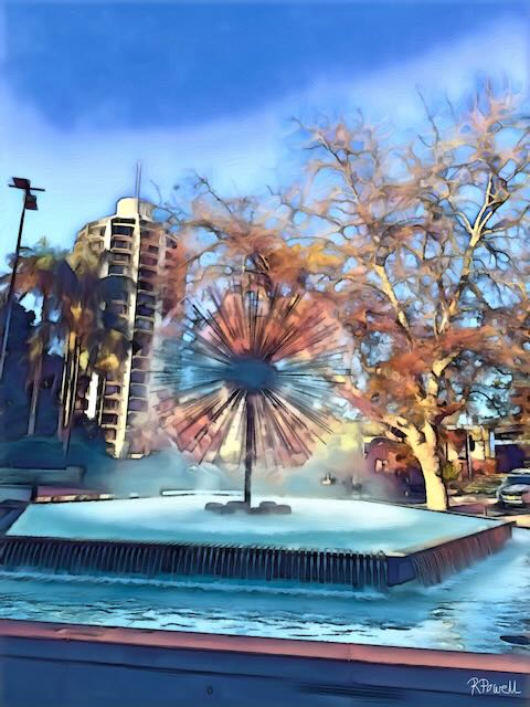 The El Alamein Fountain in Potts Point, with water spraying from a central dandelion-shaped sculpture, lit by soft golden morning sun and surrounded by autumn trees and buildings. The following images feature the same artwork in different interior settings.