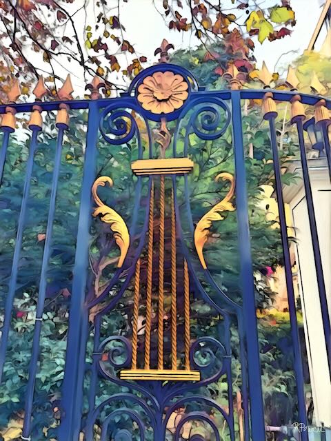 Decorative wrought iron gate with golden lyre design and floral detailing, framed by leafy greenery and autumn branches.