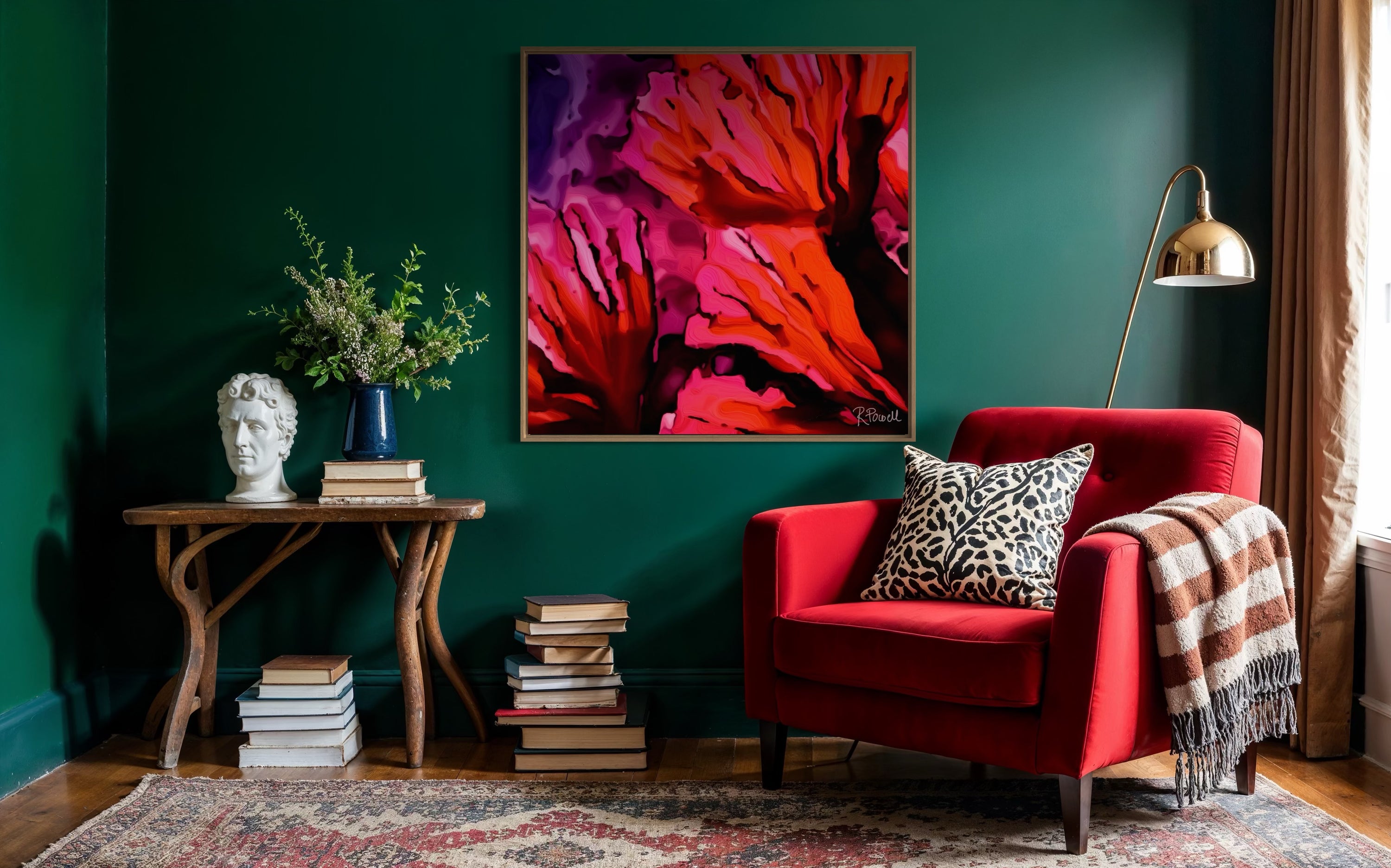 Cozy reading nook with deep green walls, a red velvet armchair, and Rosalie Powell’s Fire Bloom artwork—bold abstract petals in fiery reds and pinks. The space features a patterned rug, vintage wood table with books and a bust, a brass floor lamp, and leopard-print cushion for contrast.