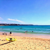 Painting of Manly Beach in the morning, with swimmers and surfers in turquoise water, golden sand in the foreground, and a bright blue sky above. The following images show the same artwork displayed in different interior settings.